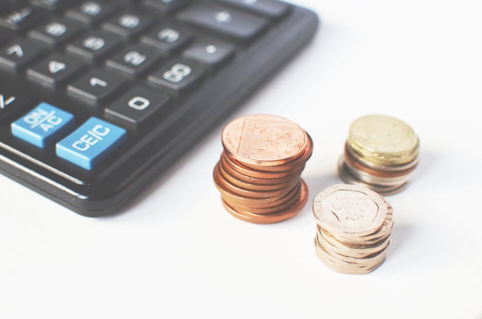 Close-up view of a black calculator with blue and white keys positioned on the left side of the image, next to three neatly stacked piles of coins in the centre and right side, consisting of copper, s