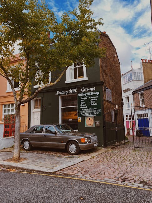 Exterior view of the Notting Hill Garage located on a narrow residential street, with a mature tree in the foreground partially obscuring a dark green building. The garage has a large sign displaying
