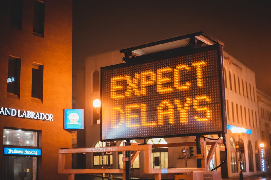A street scene at night showing a digital road sign with illuminated orange text reading 'EXPECT DELAYS' mounted on a wooden stand, with a partly visible modern brick building in the background. The s